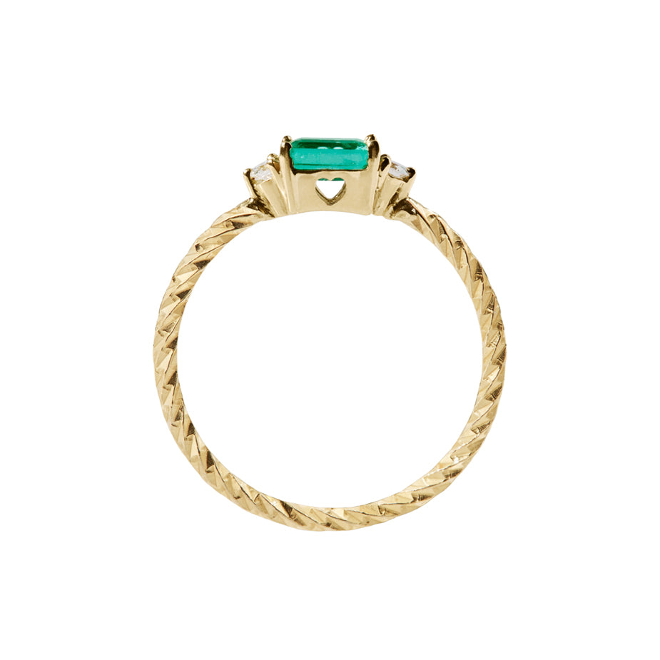 Emerald trilogy birthstone ring with two white diamond side stones on a textured gold band. Sideview showing a heart cut out on the stone setting.