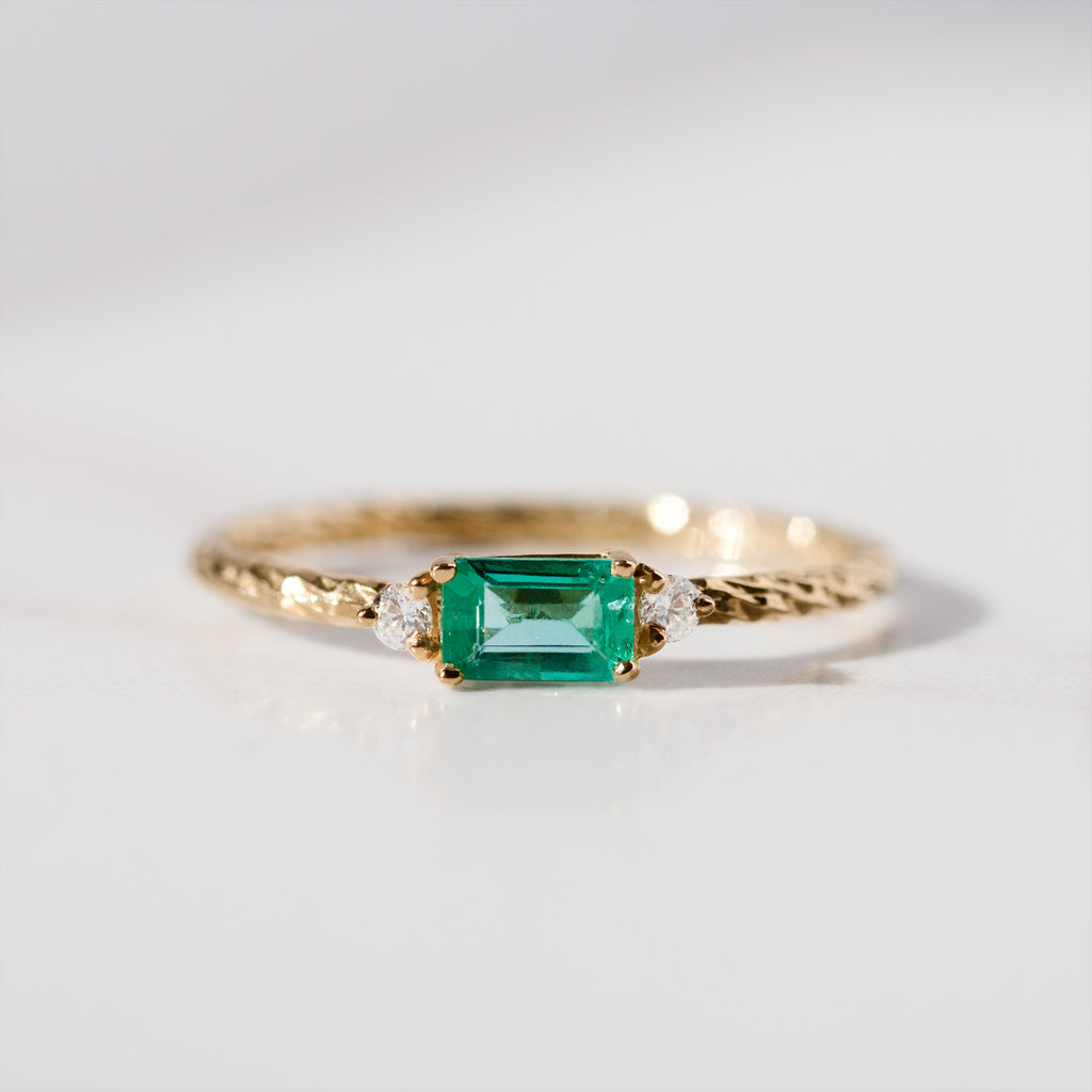 Emerald trilogy birthstone ring with two white diamond side stones on a textured gold band.